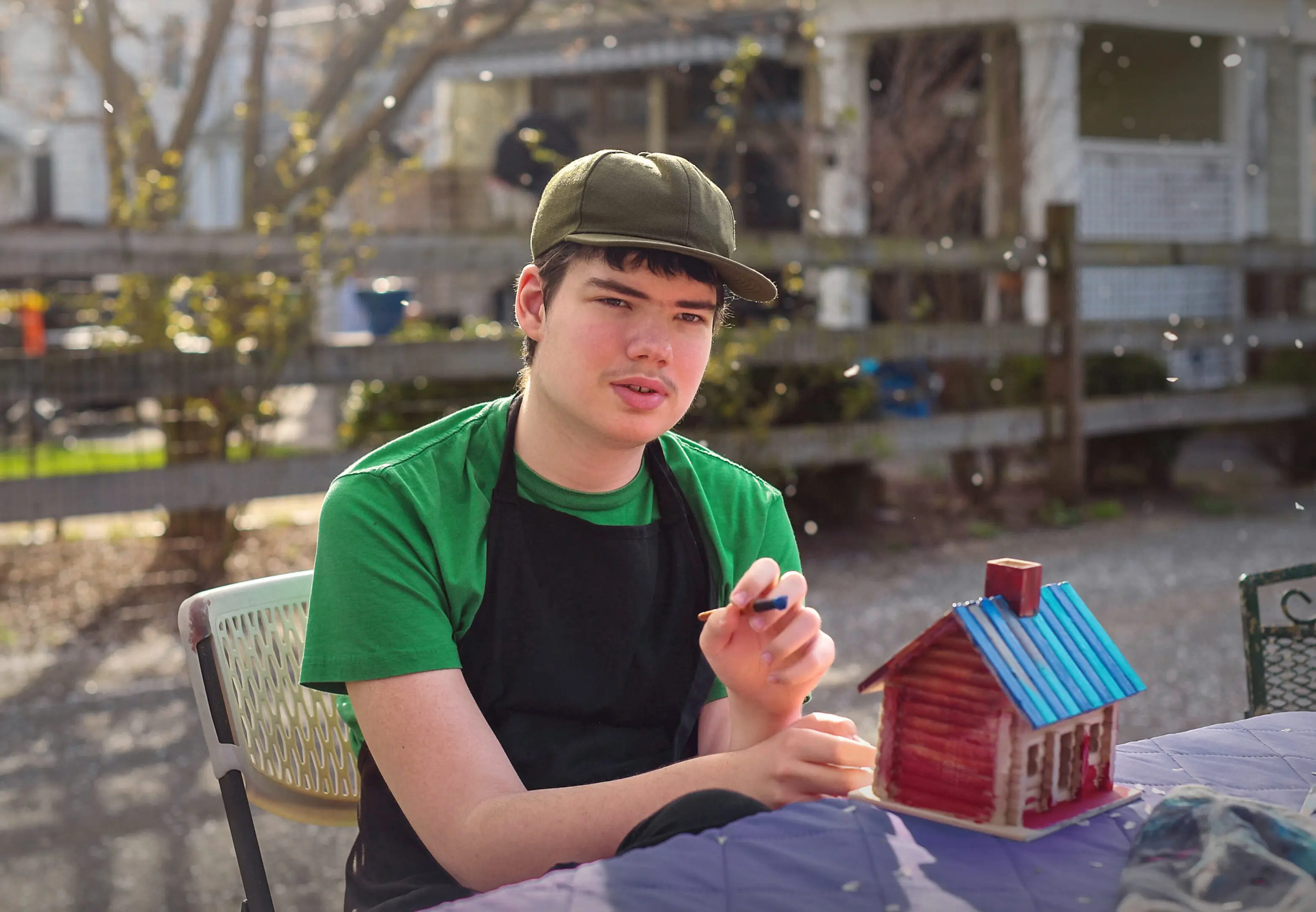 Teen boy in green shirt and cap sitting outdoors with a small house model.
