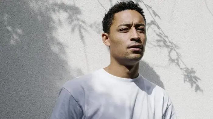 A man with curly hair wearing a white shirt stands against a light background with palm tree shadows.