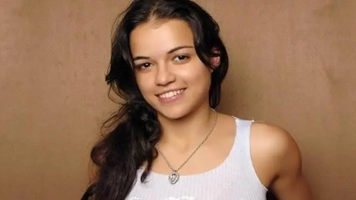 A young woman with long dark hair smiling, wearing a white tank top and a heart-shaped necklace.