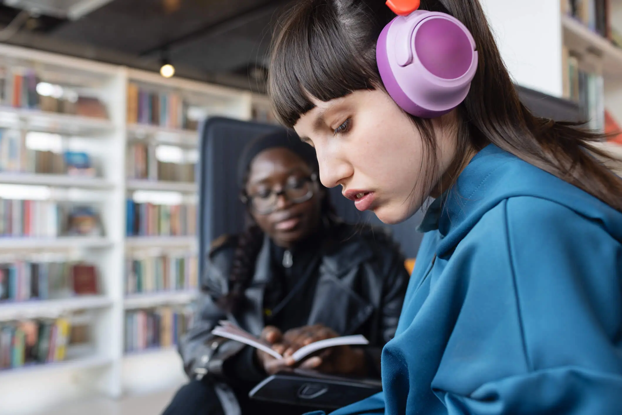 A woman wearing purple headphones and a man reading a book in a library.
