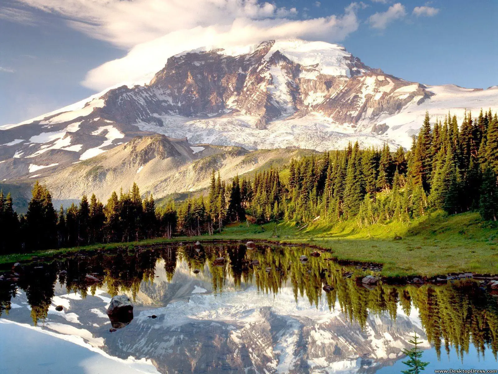 Snow-capped mountain reflected in a clear lake with green trees around.